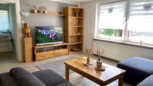 Un salon moderne avec un meuble de rangement en bois pour l’équipement audiovisuel, un téléviseur à écran plat, un canapé, une table basse et une grande fenêtre équipée de stores.