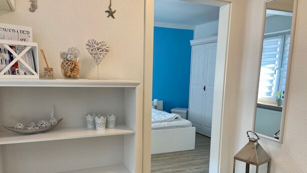 Un couloir avec une étagère blanche, un meuble en bois, une lanterne décorative et, au fond, un aperçu d’une chambre avec un mur bleu et une armoire blanche.