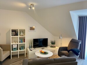 A modern living room with a flat-screen TV, a bookshelf, a dining table with fruit, a sofa, and a chair. - 7008 - An der Aue 9a - 7008 - "Domicil" - An der Aue 9a - Apartment 19 (Dahme)