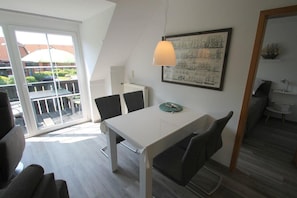 A modern, well-lit room with a dining table, chairs, and a pendant light. A large window provides a view of the outdoors. - 7008 - An der Aue 9a - 7008 - "Domicil" - An der Aue 9a - Apartment 19 (Dahme)