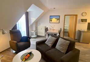 Ein modernes Wohnzimmer mit einer Couch, einem Sessel und einem Esstisch. Es gibt eine kleine Küchenzeile mit Kühlschrank und Mikrowelle. Ein Gemälde hängt an der Wand, und eine Uhr ist sichtbar.