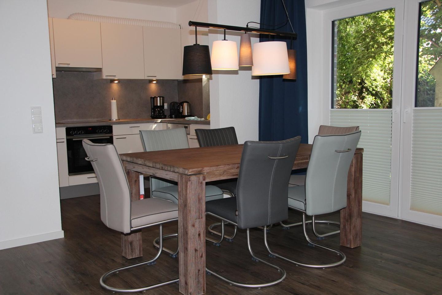A modern kitchen with white cabinets, a built-in oven, and a coffee maker. There is a dining table with six chairs and a hanging light fixture above it.