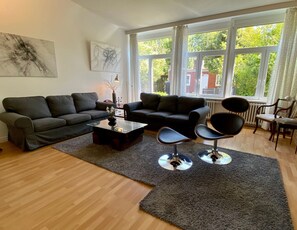 A modern living room with a sofa, coffee table, and two chairs. Large windows provide natural light and a view of greenery outside.