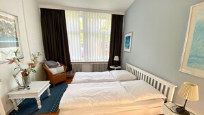 A hotel room with two beds, a small table, a chair, a radiator, and a window with curtains.
