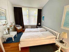 Habitación de hotel con dos camas, una mesita pequeña, una silla, un radiador y una ventana con cortinas.
