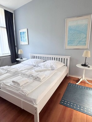 Ein Hotelzimmer mit einem Bett, zwei Nachttischen, einem Fenster mit Vorhängen, einem Heizkörper, einem Teppich und zwei gerahmten Bildern an der Wand.