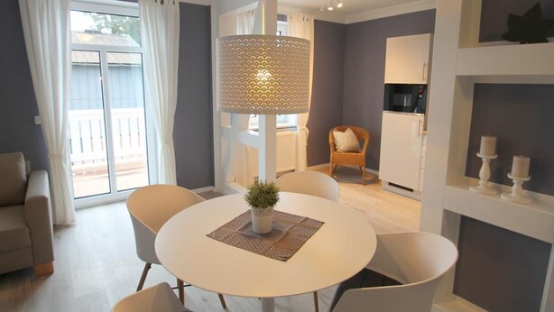 A modern living room with a round dining table, white chairs, a hanging light fixture, and a view of a balcony through large windows.