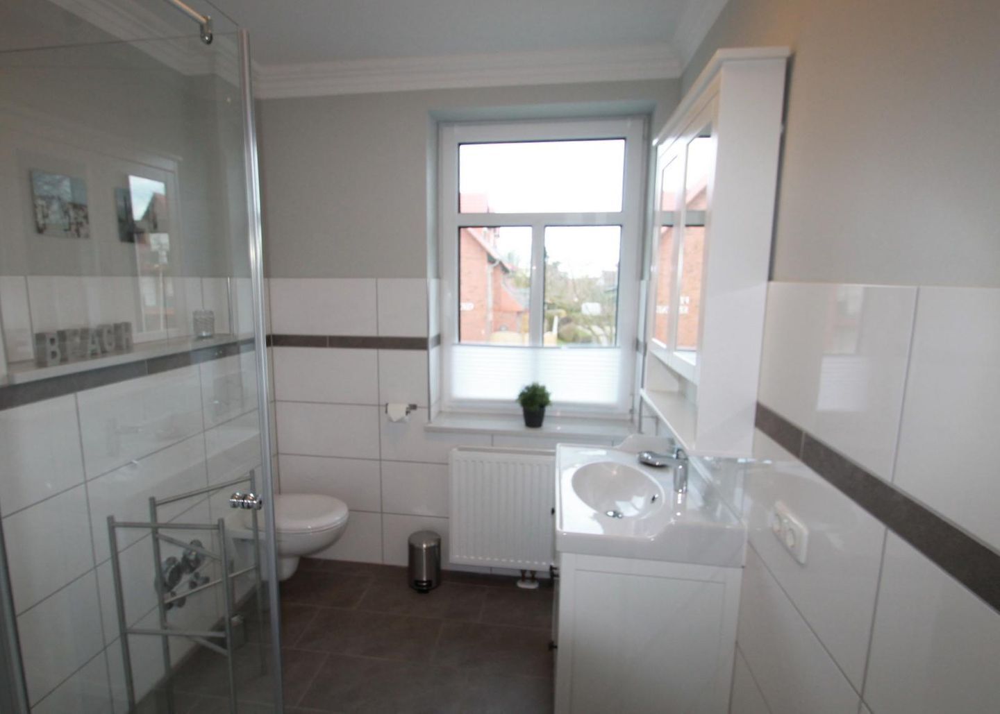 Et badeværelse med glasbrusekabine, toilet, vask og et vindue med udsigt til bygninger.