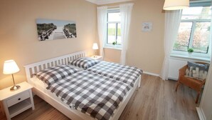 A bedroom with two beds, a bedside table with a lamp, a window with curtains, and a painting on the wall. - Beach shell - Alte Da(h)me - New 1024 - Beach shell - Die Alte Dahme - New (Dahme)