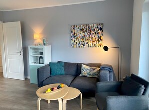 A modern living room with a grey sofa, a small round table with fruit, a bookshelf, and a colorful abstract painting on the wall.