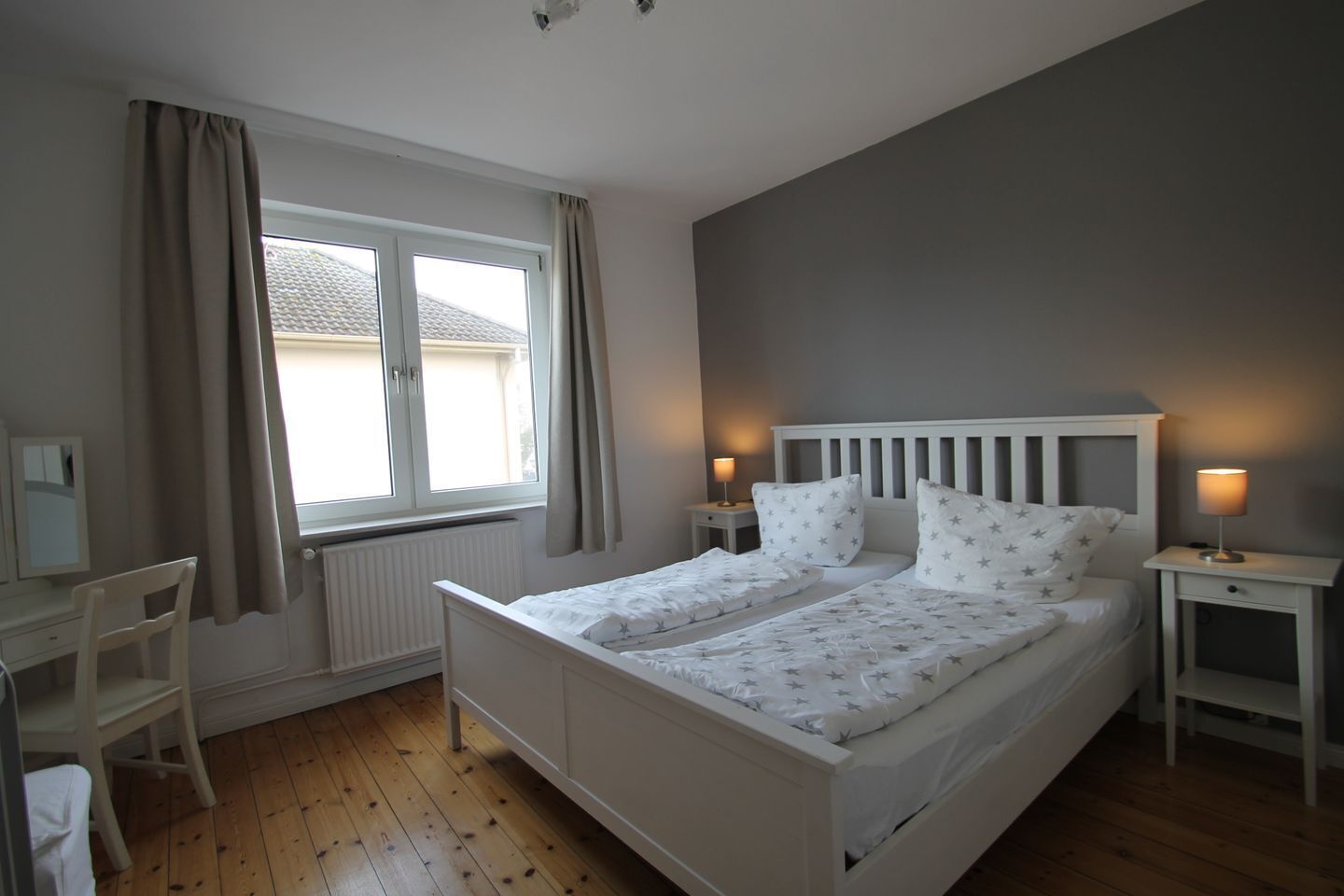 A bedroom with a double bed, two bedside tables with lamps, a desk with a chair, and a window with curtains.