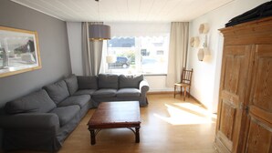 Un salon avec un canapé d’angle gris, une table basse en bois, un meuble en bois et une fenêtre avec des rideaux.