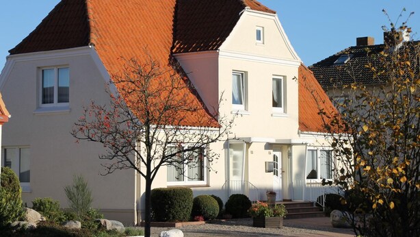 Une maison à deux étages, avec un toit recouvert de tuiles rouges, des murs extérieurs blancs et un petit porche à l’avant.