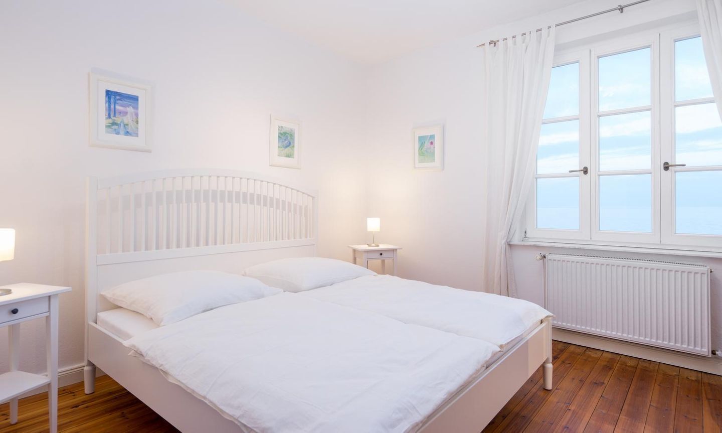 A white bedroom with a bed, a nightstand, a window with curtains, and a radiator.