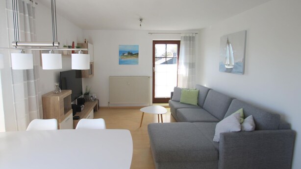 A modern living room with a grey sofa, a white table, and a kitchen area with a radiator and a painting on the wall.