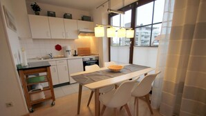 A modern kitchen with white cabinets, a dining table with chairs, a wooden sideboard, and a large window with curtains.