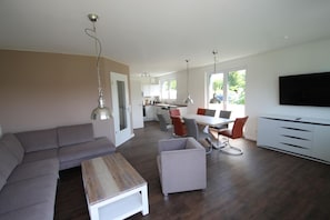 A modern living room with a sofa, a coffee table, a dining area with chairs, a kitchen in the background, and a TV mounted on the wall.