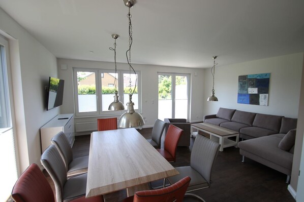 Una sala de estar moderna con una mesa de comedor, sofá y sillas. Hay ventanas grandes y un televisor de pantalla plana montado en la pared.
