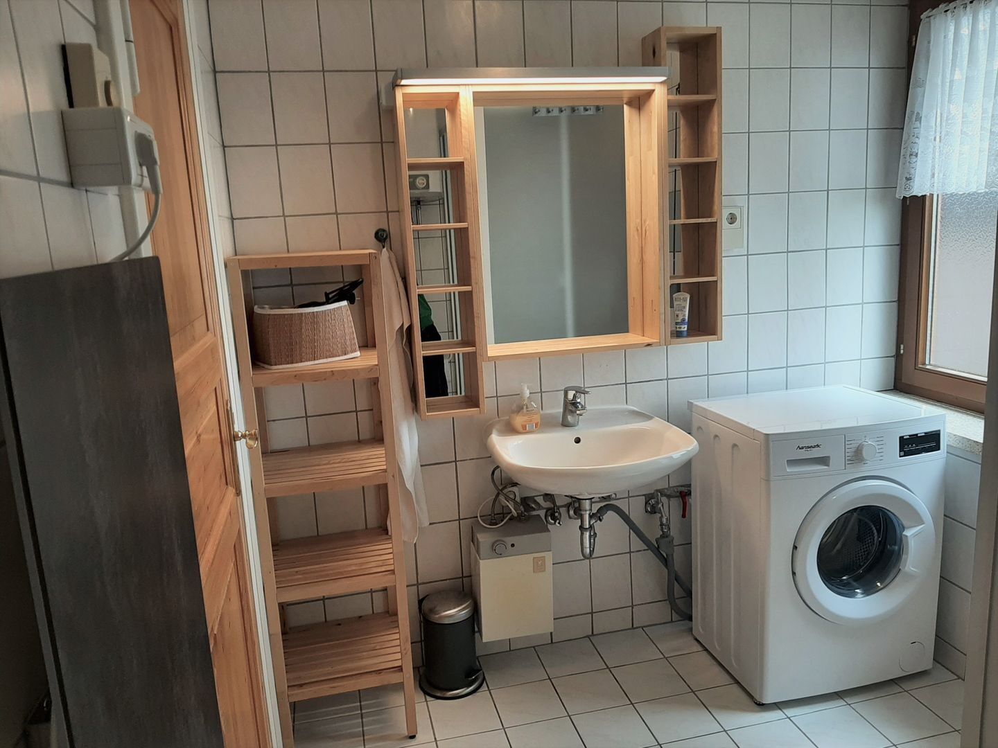 Ein kleines Badezimmer mit Waschbecken, Spiegel, Holzregalen, einer Waschmaschine und einem Mülleimer.