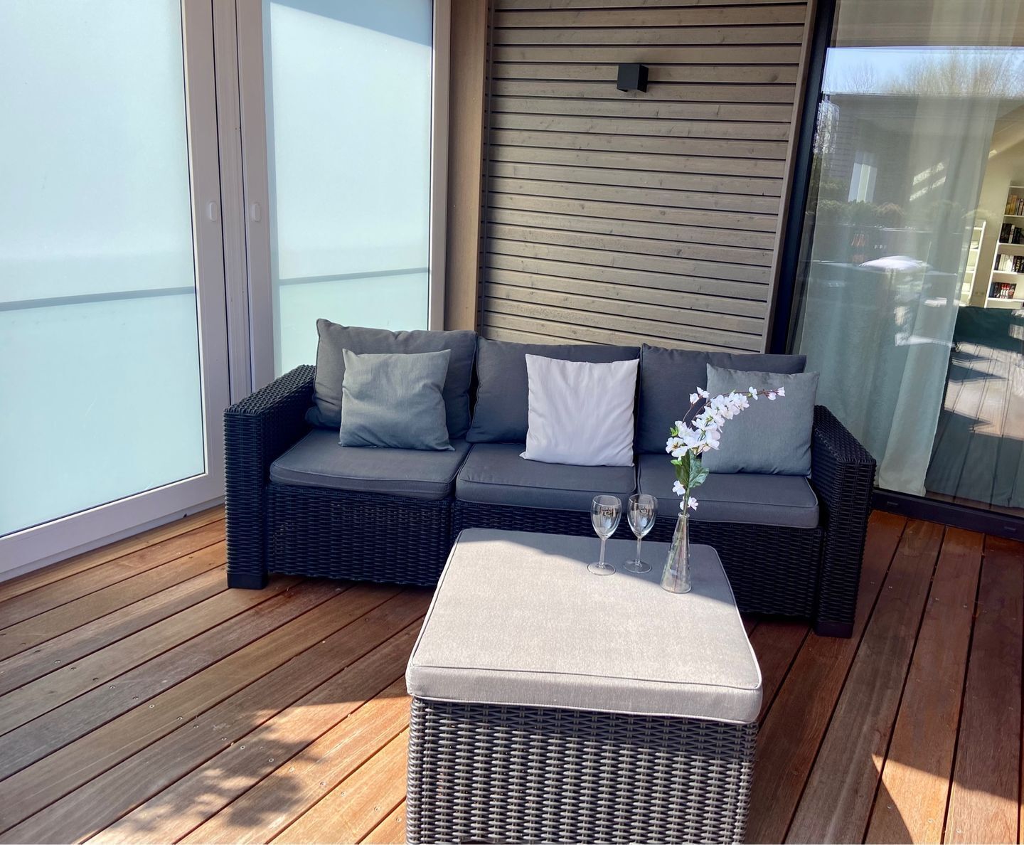 A balcony with a wicker sofa, cushions, and a small table with a vase of flowers.