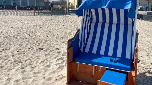 Une chaise de plage recouverte d’une toile bleue et blanche à rayures, placée sur une plage de sable, avec un bâtiment portant l’inscription « DUS RESTAURANT & CAFE AM MEER » en arrière-plan.