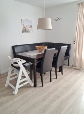 Un comedor con una mesa de madera oscura, cuatro sillas negras, una trona blanca, una cesta tejida, una lámpara colgante y un cuadro en la pared.