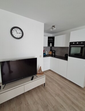Une cuisine moderne avec des meubles blancs, un four encastré et un téléviseur à écran plat posé sur une console blanche.