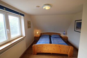 A small, cozy room with a wooden bed, a nightstand with a lamp, a window with blinds, and a ceiling light.