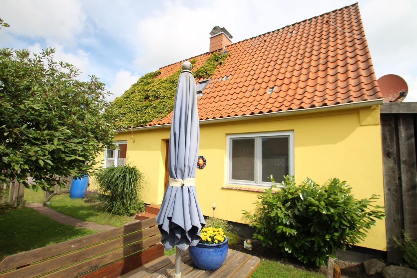 Une maison jaune avec un toit recouvert de tuiles rouges, un parasol bleu et une plante en pot ornée de fleurs jaunes.