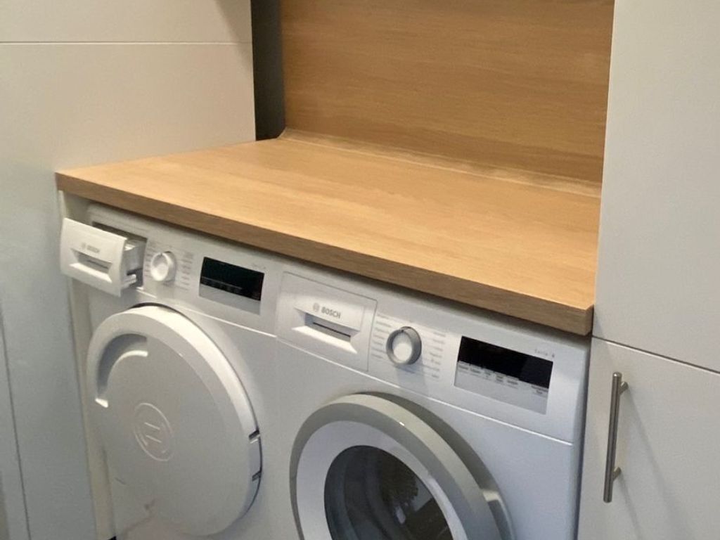 A front view of a Bosch washing machine in a laundry room with a wooden countertop and a grey cabinet.