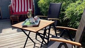 Une terrasse en bois avec une table et deux chaises, un parasol rouge et blanc à rayures, et une végétation luxuriante.