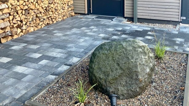 Un bâtiment moderne avec une porte sombre, un mur de pierre et un gros rocher au premier plan.