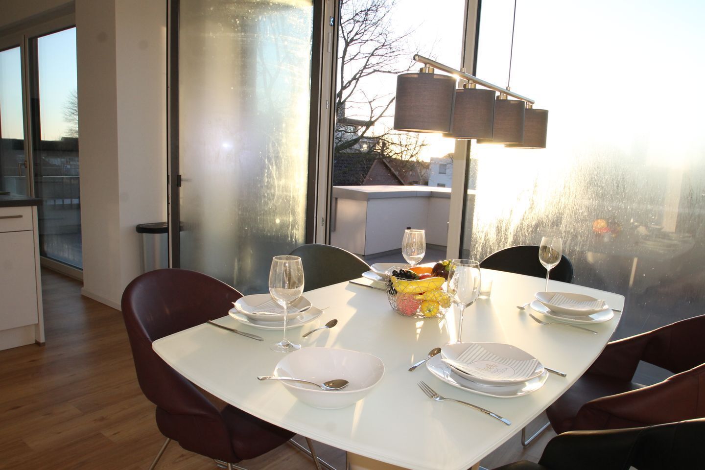 Une table à manger dressée avec de l’argenterie, des assiettes blanches et un panier de fruits, offrant une vue sur la ville à travers de grandes fenêtres.