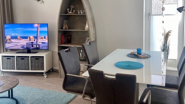 A modern living room with a television, a dining table with chairs, a bookshelf, and a large window.