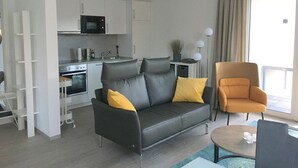 A modern living room with a grey sofa, a yellow pillow, and an orange armchair. There is a kitchen area with white cabinets and a microwave.