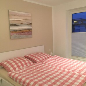 A bedroom with a bed, checkered bedding, a painting on the wall, and a window with a view of the outside.