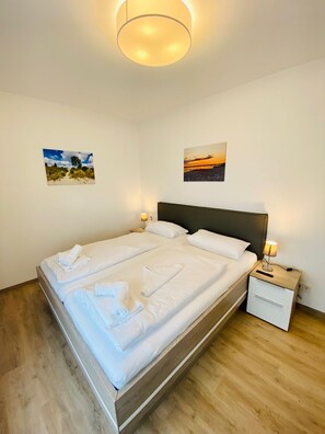 Un lit double avec du linge de lit et des serviettes blanches, deux tables de chevet avec des lampes, un plancher en bois et deux tableaux accrochés au mur.