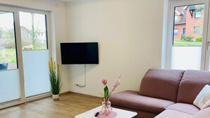 A modern living room with a pink sofa, a flat-screen TV mounted on the wall, a small white table with a vase of flowers, and a large window providing a view of the outside. - 2803 - Strandliebe - Apartment Strandliebe (Dahme)