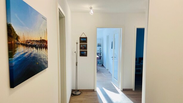 A hallway with a wooden floor, a painting of a waterfront scene, and a floor lamp. - 2803 - Strandliebe - Apartment Strandliebe (Dahme)