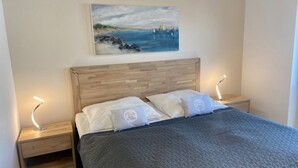 Een slaapkamer met een houten hoofdbord, een bed met een grijs geborduurd sprei, twee nachtkastjes met lampen en een schilderij van een kustlandschap aan de muur.