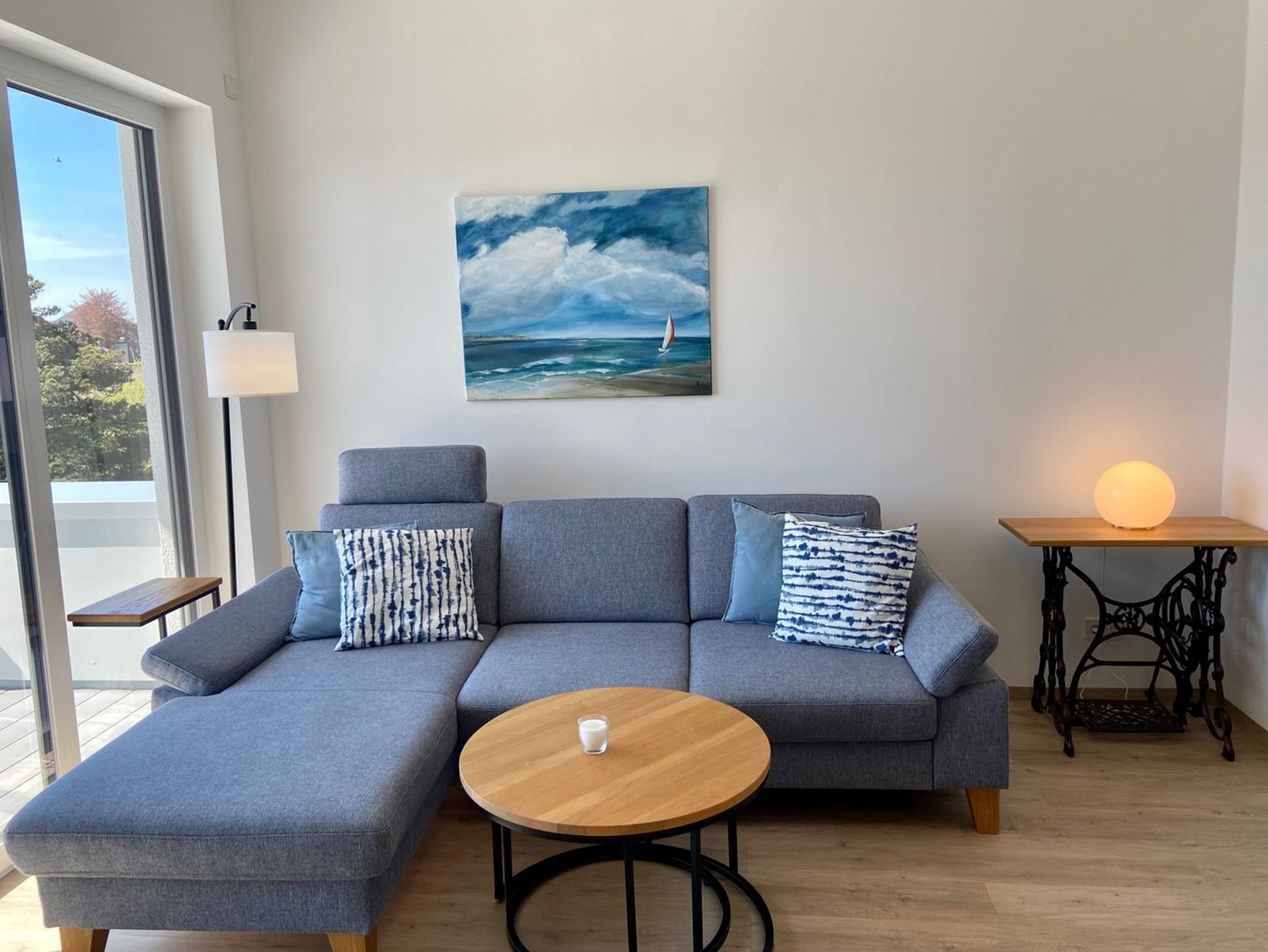 Ein modernes Wohnzimmer mit einem grauen Ecksofa, einem runden Couchtisch aus Holz und einem Gemälde mit einem Segelboot auf dem Meer.