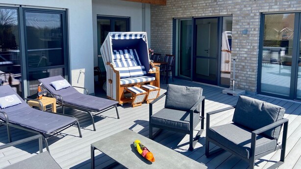 Een moderne terrasruimte met ligbedden, een rieten stoel en een tafel met fruit.