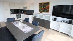 Een moderne keuken met witte kastjes, een zwarte kookplaat en een magnetron. Er is een eethoek met een tafel, stoelen en een vaas met bloemen. Een schilderij hangt aan de muur, en een televisie is bevestigd boven een witte plank met speakers.