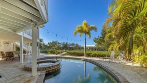 A swimming pool with a curved edge, surrounded by a brick patio. There is a covered seating area with chairs and a table, and a palm tree in the background. - Pool home on Bocilla Lagoon (Placida)