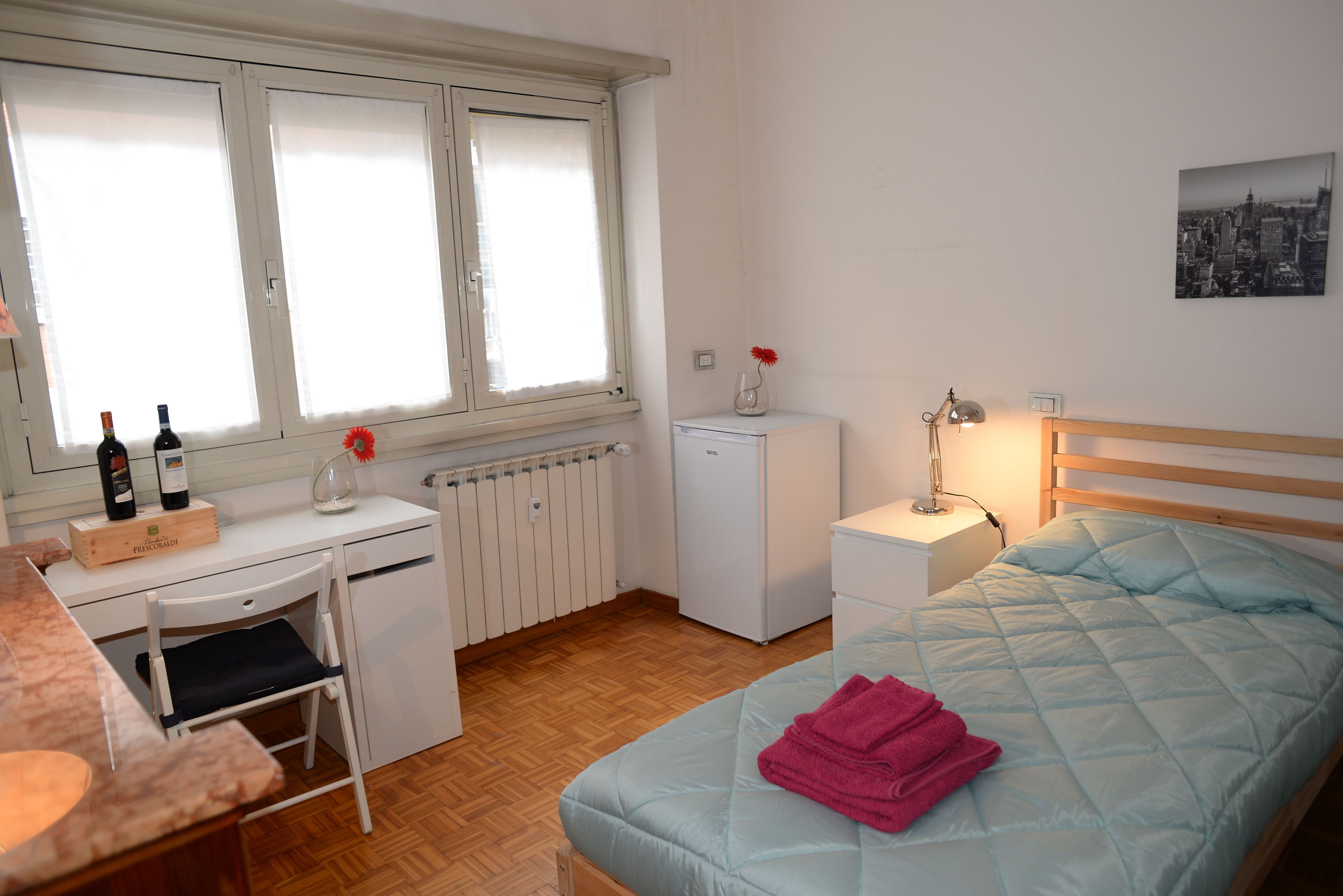 Une chambre à coucher comprenant un lit, un bureau, une chaise, un radiateur, un petit réfrigérateur et une fenêtre avec des stores.