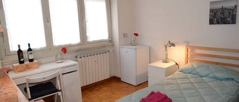 Ein Schlafzimmer mit Bett, Schreibtisch, Stuhl, Heizkörper, kleinem Kühlschrank und einem Fenster mit Jalousien.