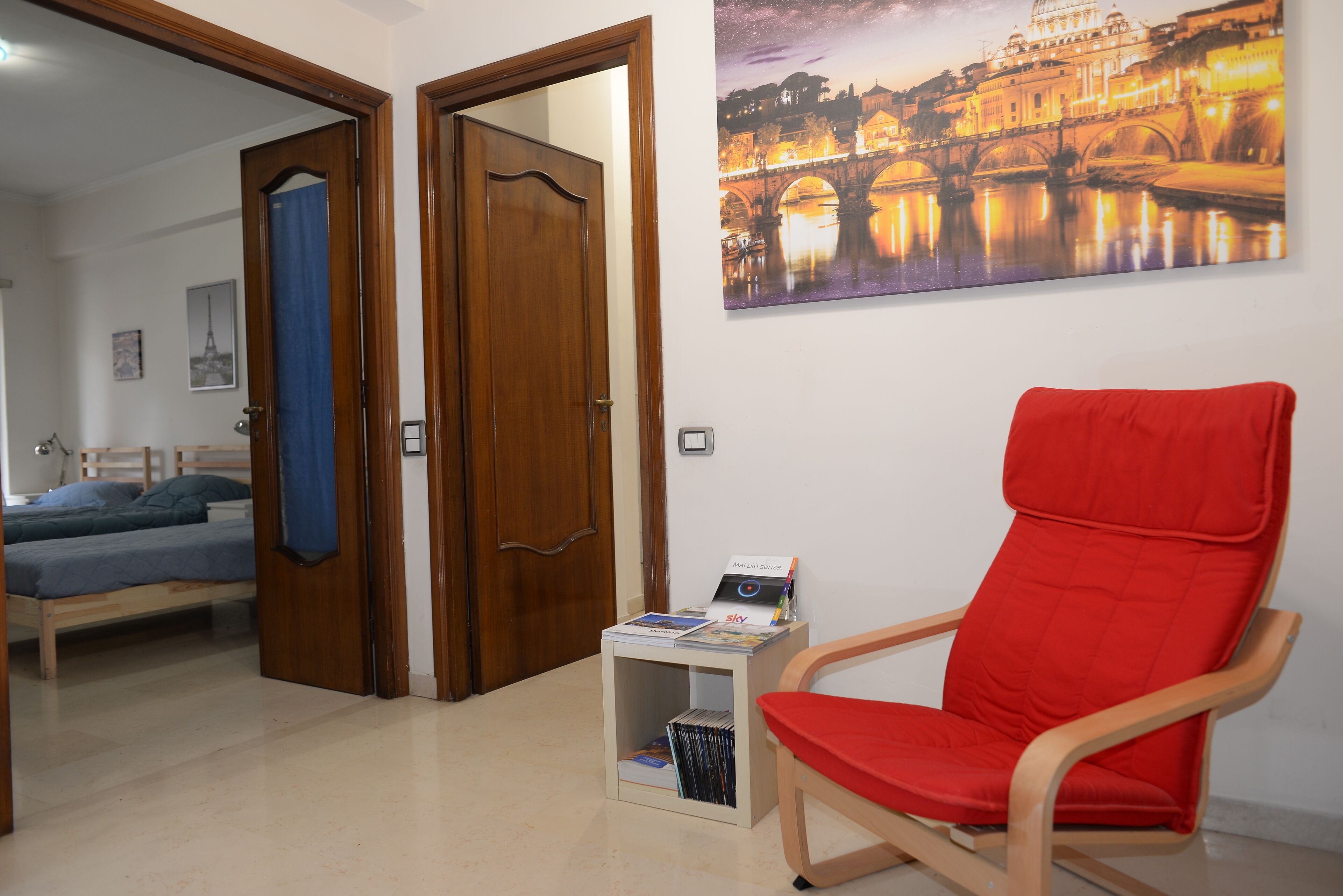 Une pièce comprenant un lit, un fauteuil rouge, une petite table avec des magazines et un tableau représentant un pont.