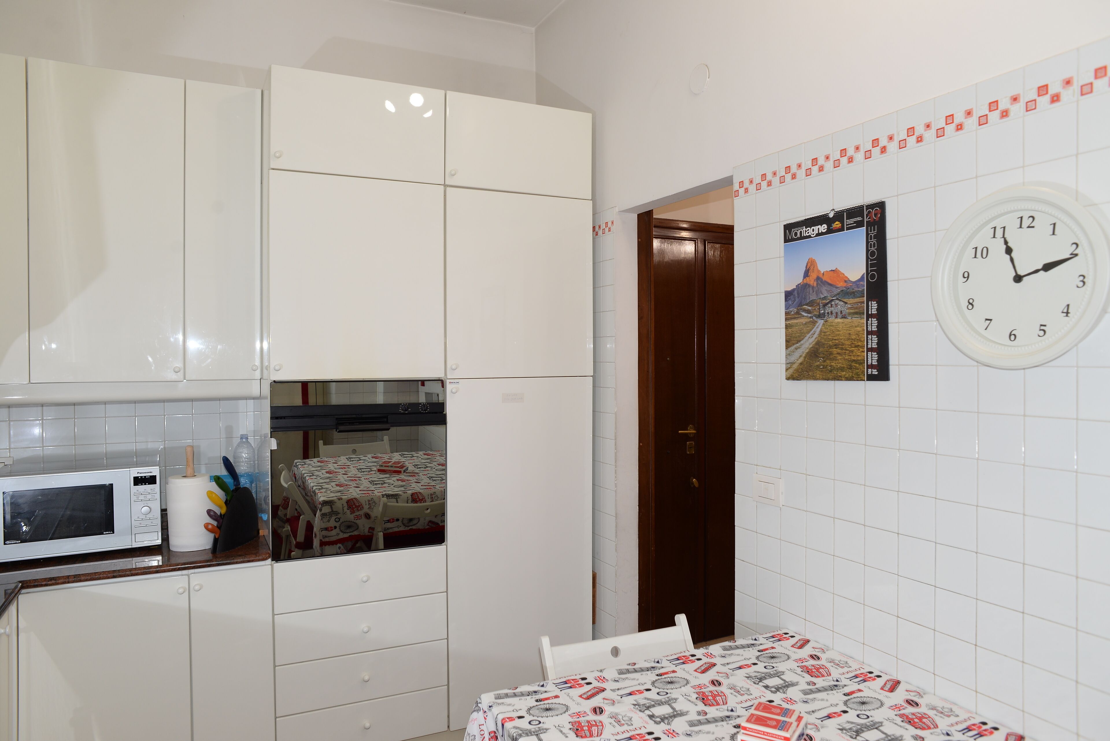 Une cuisine avec des meubles blancs, un four à micro-ondes et une table recouverte d’une nappe à motifs.