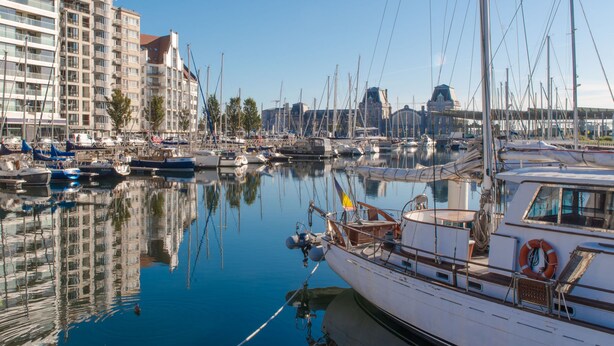 Een jachthaven met aangemeerde boten, een groot gebouw met meerdere verdiepingen, en een heldere blauwe lucht.