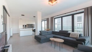 Een moderne woonkamer met een grijze hoekbank, een klein rond salontafeltje en een flatscreen-tv. Achtergrond toont een keuken met witte kasten en een centraal keukenblok.